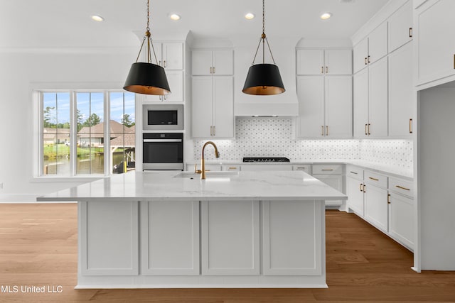 kitchen with oven, a center island with sink, white cabinetry, light stone countertops, and light wood-type flooring