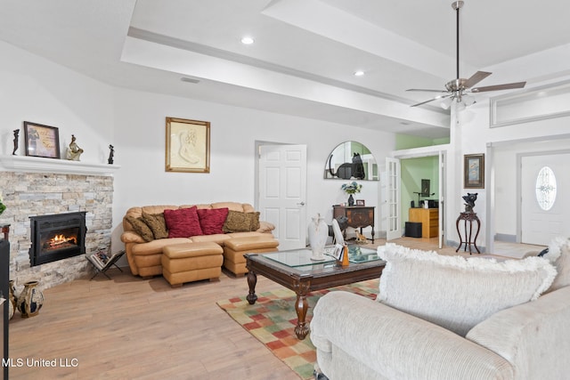 living room with a tray ceiling, a stone fireplace, ceiling fan, and light hardwood / wood-style floors