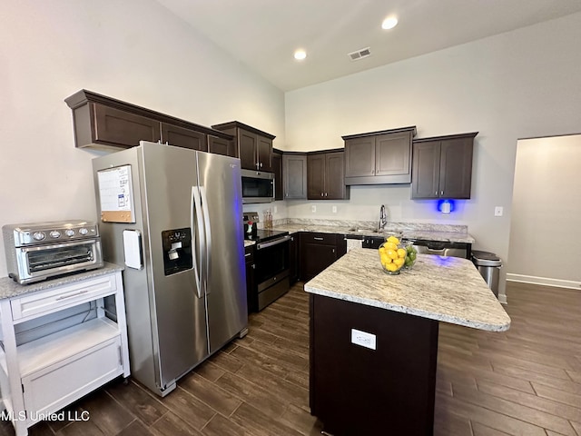 kitchen with stainless steel appliances, dark wood-style floors, dark brown cabinets, light stone countertops, and a center island