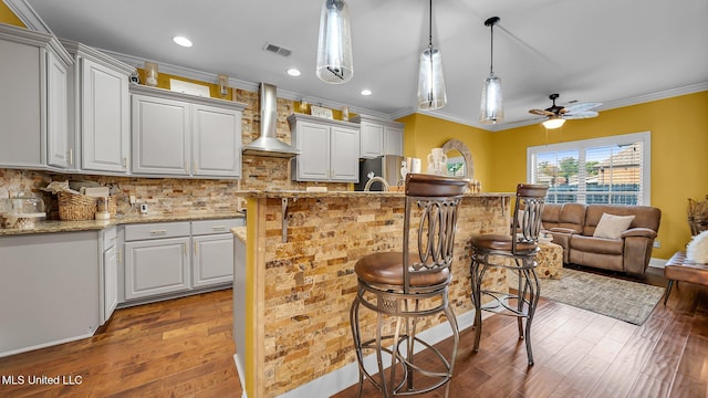 kitchen with light stone countertops, stainless steel fridge, hardwood / wood-style floors, ceiling fan, and wall chimney exhaust hood