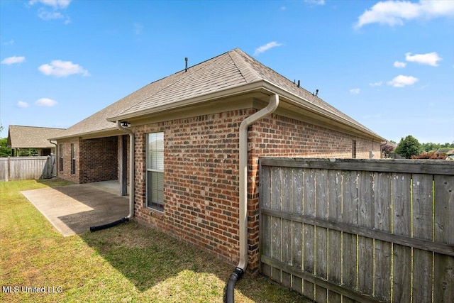 view of side of home with fence, a shingled roof, brick siding, a patio area, and a lawn