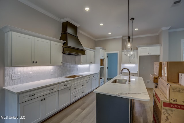 kitchen featuring a kitchen island with sink, custom range hood, sink, decorative light fixtures, and light wood-type flooring