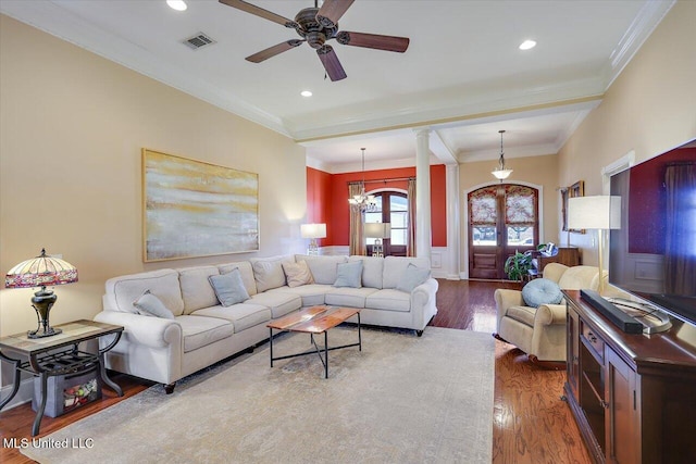 living room featuring ornate columns, ceiling fan with notable chandelier, ornamental molding, dark wood-type flooring, and french doors