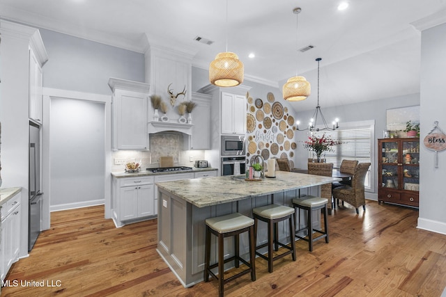 kitchen featuring appliances with stainless steel finishes, light stone counters, white cabinets, a center island with sink, and decorative light fixtures