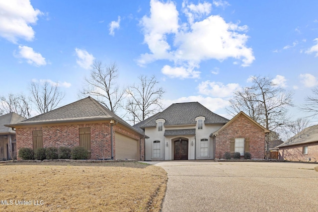 french country style house with an attached garage, brick siding, concrete driveway, roof with shingles, and stucco siding