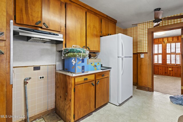 kitchen featuring under cabinet range hood, wallpapered walls, freestanding refrigerator, brown cabinetry, and wainscoting