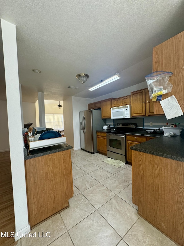 kitchen featuring light tile patterned floors, a peninsula, stainless steel appliances, dark countertops, and brown cabinets