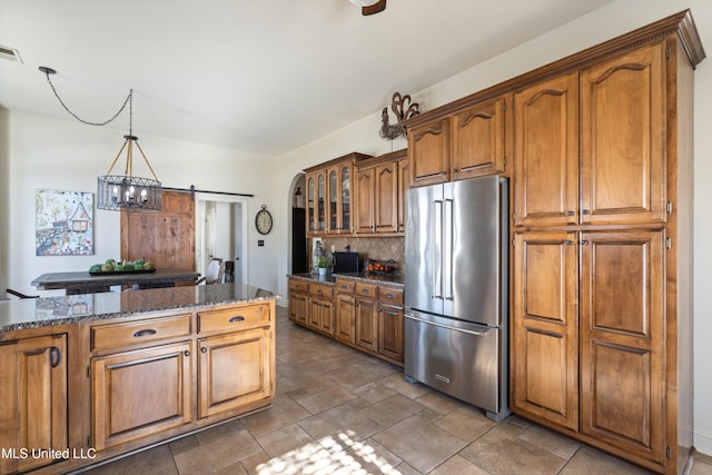 kitchen with decorative backsplash, high end fridge, dark stone counters, a barn door, and decorative light fixtures