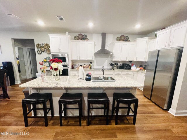 kitchen featuring wall chimney range hood, a kitchen island with sink, white cabinetry, a kitchen bar, and appliances with stainless steel finishes