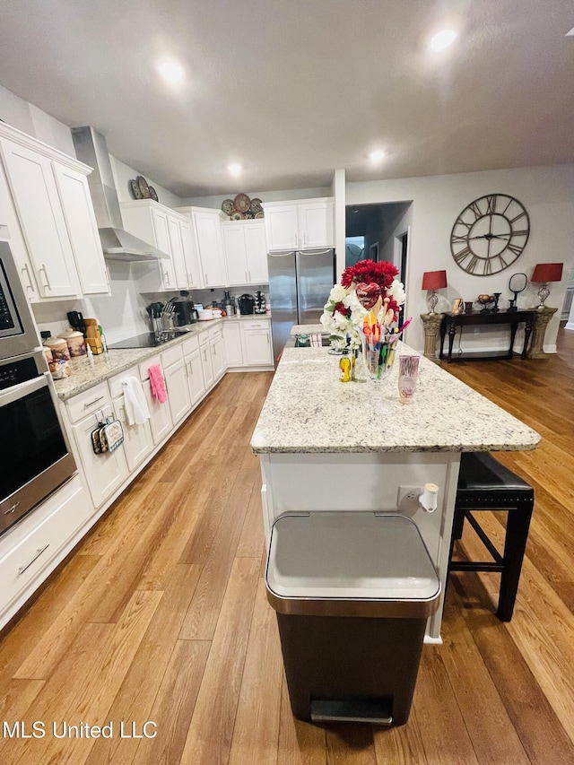 kitchen featuring white cabinets, light wood-type flooring, a kitchen island, wall chimney exhaust hood, and appliances with stainless steel finishes