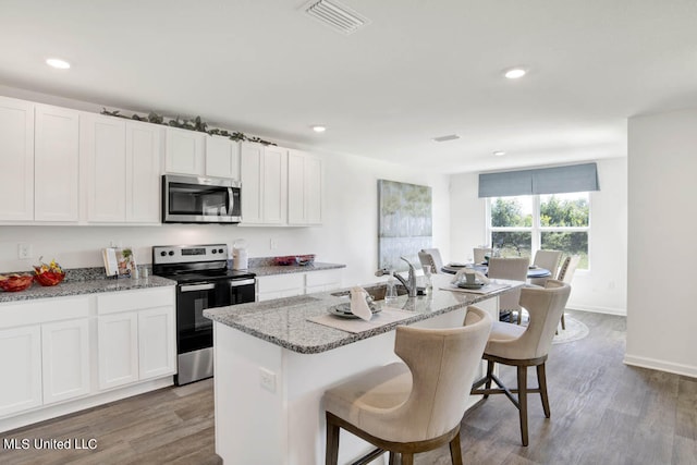 kitchen featuring stainless steel appliances, white cabinets, dark wood-type flooring, light stone counters, and a center island with sink