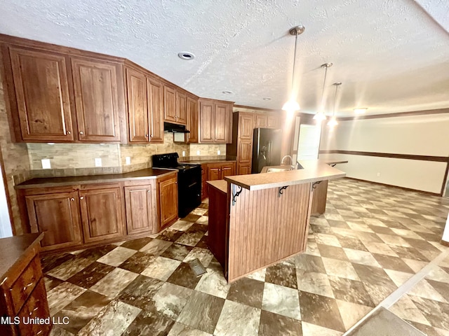 kitchen featuring under cabinet range hood, black electric range, a kitchen island with sink, brown cabinetry, and stainless steel fridge with ice dispenser