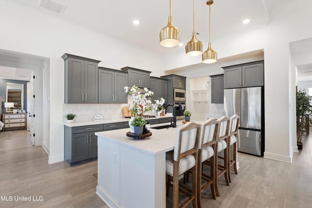 kitchen featuring stainless steel appliances, a center island with sink, decorative backsplash, pendant lighting, and light hardwood / wood-style flooring