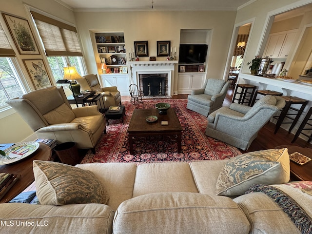 living area featuring built in shelves, plenty of natural light, wood finished floors, a fireplace with flush hearth, and crown molding