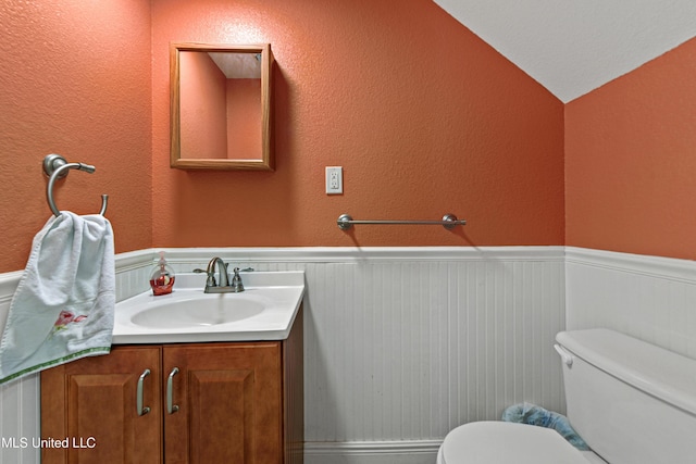 bathroom with a textured wall, toilet, a wainscoted wall, vanity, and vaulted ceiling