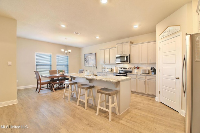 kitchen featuring appliances with stainless steel finishes, a kitchen island with sink, light stone counters, light hardwood / wood-style floors, and decorative light fixtures