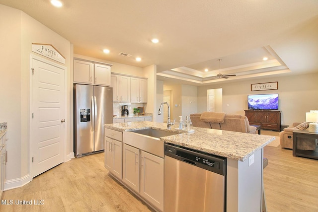 kitchen featuring sink, light wood-type flooring, a tray ceiling, stainless steel appliances, and a kitchen island with sink