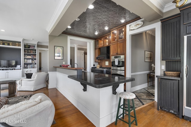 kitchen with exhaust hood, dark hardwood / wood-style flooring, a kitchen bar, ornamental molding, and built in shelves