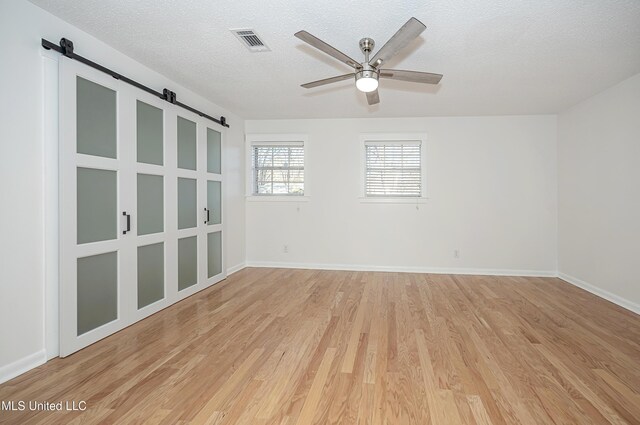 empty room featuring a textured ceiling, a barn door, a ceiling fan, visible vents, and light wood-style floors