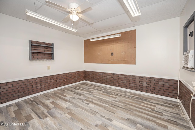 empty room featuring a paneled ceiling, a ceiling fan, wainscoting, brick wall, and wood finished floors