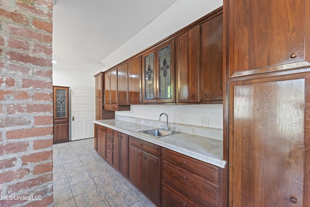 kitchen with tile counters, glass insert cabinets, a sink, and brick wall