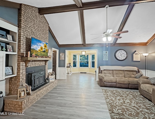 living area featuring a ceiling fan, wainscoting, wood finished floors, beamed ceiling, and a brick fireplace