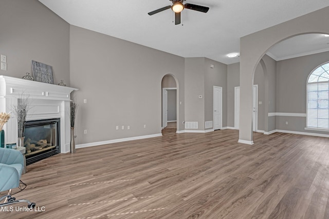 unfurnished living room featuring light wood finished floors, arched walkways, baseboards, a glass covered fireplace, and ceiling fan