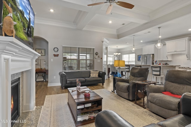 living room with beam ceiling, ceiling fan with notable chandelier, a warm lit fireplace, wood finished floors, and crown molding