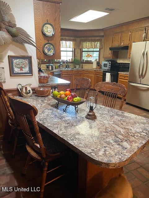 kitchen featuring under cabinet range hood, a peninsula, stainless steel appliances, and brown cabinetry