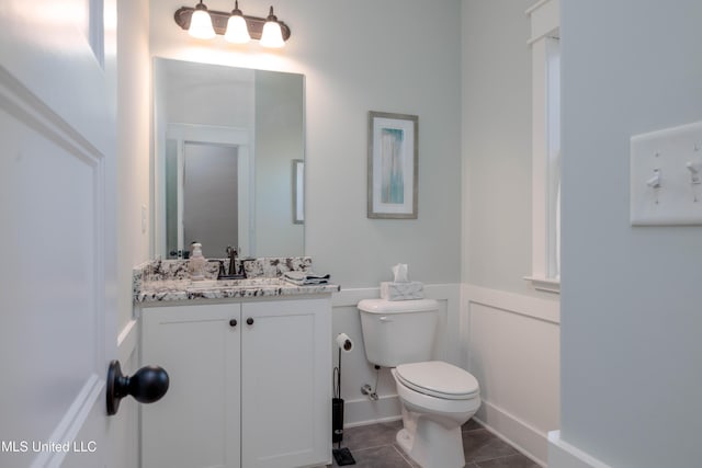 half bathroom featuring vanity, tile patterned flooring, wainscoting, a decorative wall, and toilet