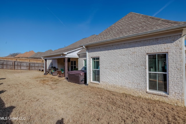 rear view of property featuring fence, brick siding, roof with shingles, and a hot tub