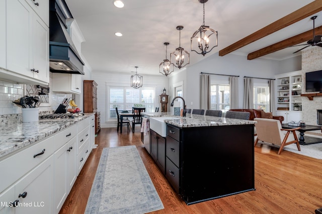 kitchen featuring stainless steel gas cooktop, custom exhaust hood, light wood-style flooring, a sink, and a stone fireplace