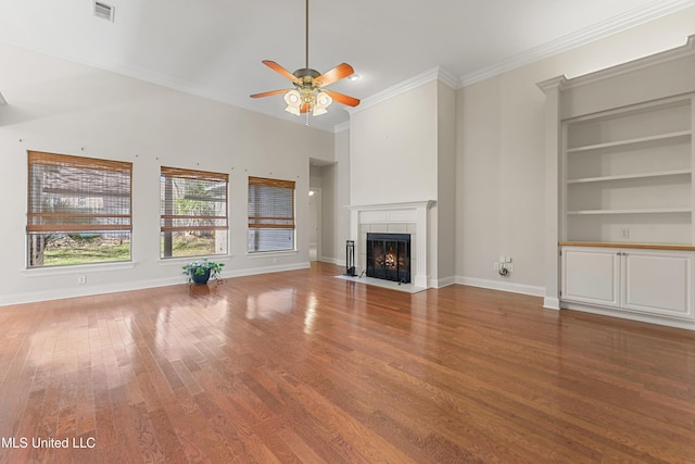 unfurnished living room featuring hardwood / wood-style floors, built in shelves, ceiling fan, a tile fireplace, and crown molding