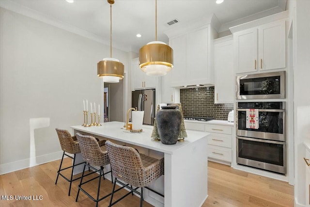 kitchen with appliances with stainless steel finishes, light hardwood / wood-style flooring, white cabinetry, hanging light fixtures, and an island with sink