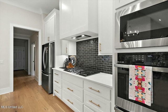 kitchen featuring wall chimney range hood, light wood-type flooring, appliances with stainless steel finishes, tasteful backsplash, and white cabinetry