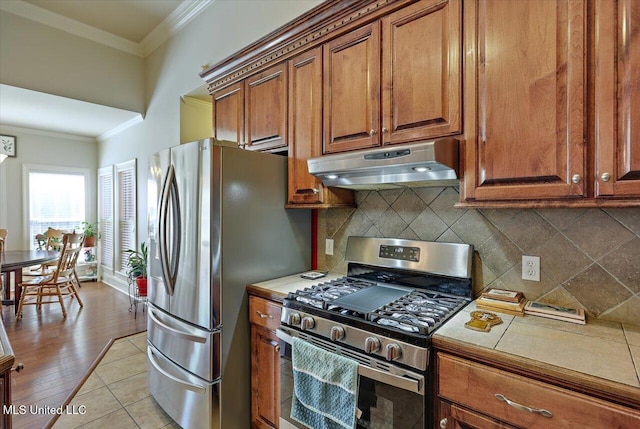 kitchen with tile countertops, ornamental molding, brown cabinets, stainless steel appliances, and under cabinet range hood