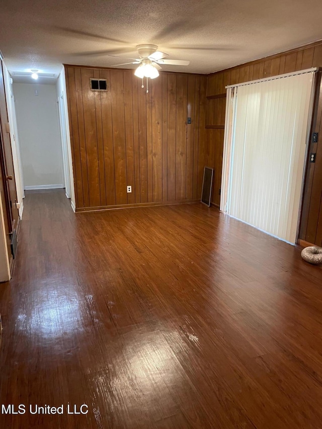 empty room featuring dark wood finished floors, a ceiling fan, wood walls, baseboards, and a textured ceiling
