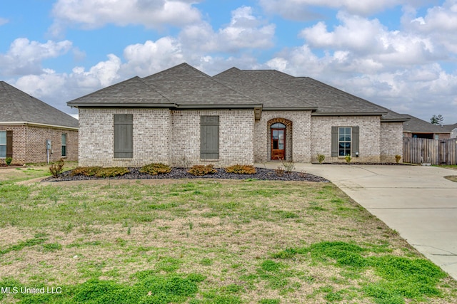 french country home with a front yard, brick siding, fence, and roof with shingles