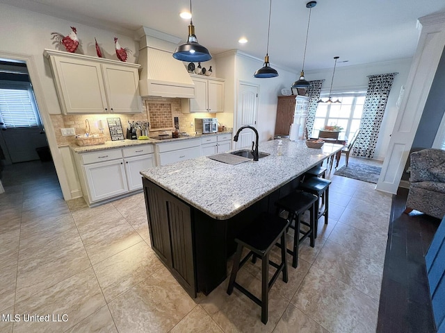 kitchen featuring sink, custom exhaust hood, decorative light fixtures, a kitchen island with sink, and white cabinets