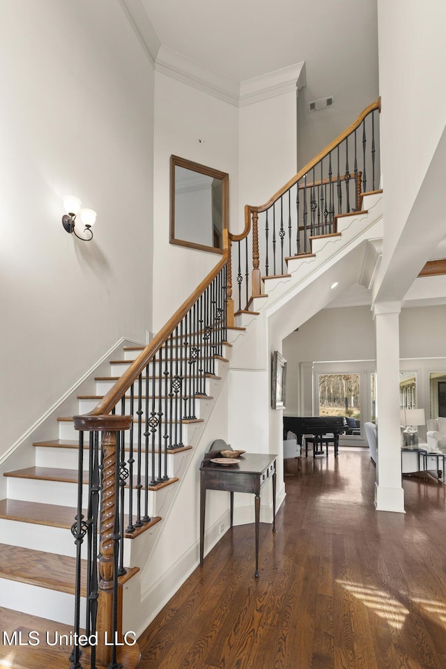 stairs with visible vents, a towering ceiling, ornamental molding, wood finished floors, and ornate columns