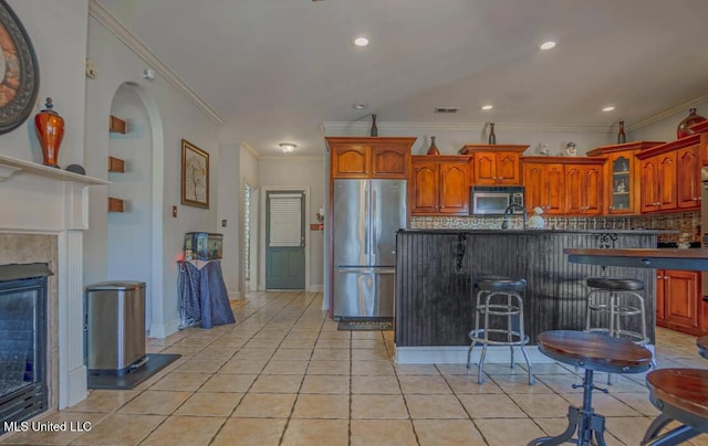 kitchen with a fireplace, light tile patterned flooring, a kitchen breakfast bar, crown molding, and appliances with stainless steel finishes