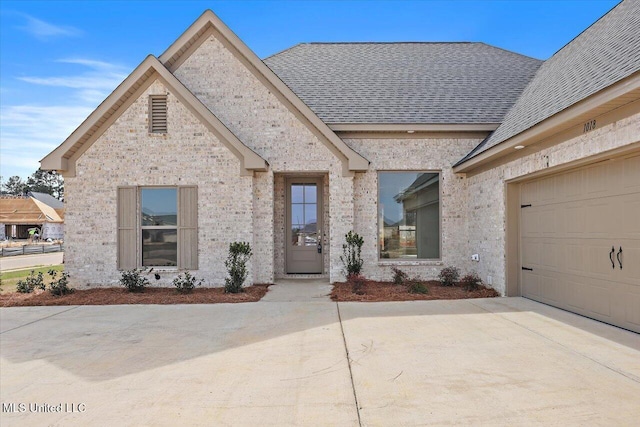 french country home with brick siding, an attached garage, driveway, and roof with shingles