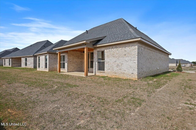 back of house featuring roof with shingles, brick siding, a patio, and a lawn