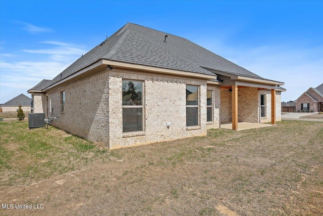 back of house with central air condition unit, brick siding, a yard, roof with shingles, and a patio area