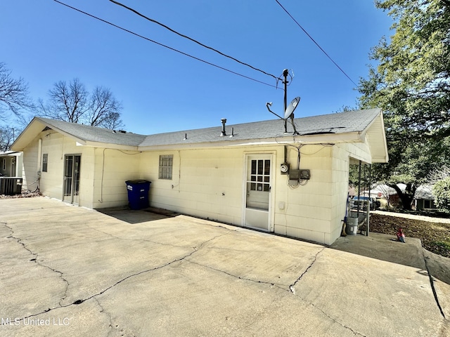 rear view of house with a shingled roof, central AC unit, concrete driveway, a patio, and concrete block siding