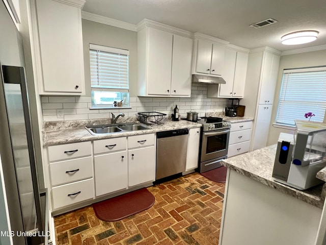 kitchen with appliances with stainless steel finishes, a sink, under cabinet range hood, brick floors, and ornamental molding