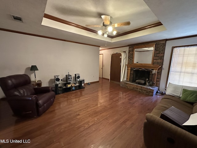 living room with dark wood-type flooring, a brick fireplace, ceiling fan, ornamental molding, and a tray ceiling