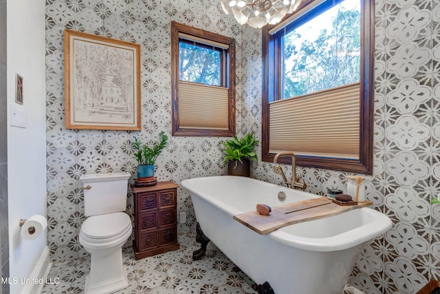 bathroom with wallpapered walls, a freestanding bath, a notable chandelier, tile patterned floors, and toilet