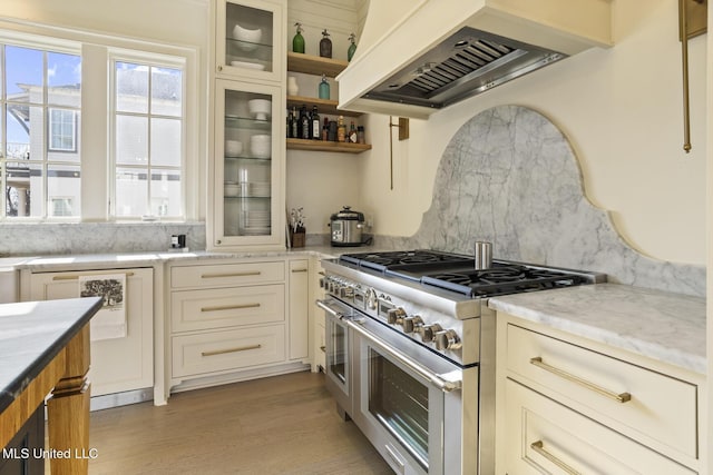 kitchen featuring premium range hood, range with two ovens, light stone countertops, light hardwood / wood-style floors, and decorative backsplash