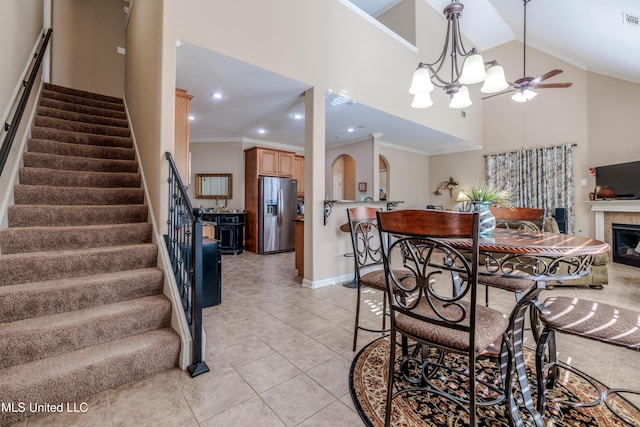 dining space with ceiling fan with notable chandelier, a fireplace, ornamental molding, high vaulted ceiling, and light tile patterned floors